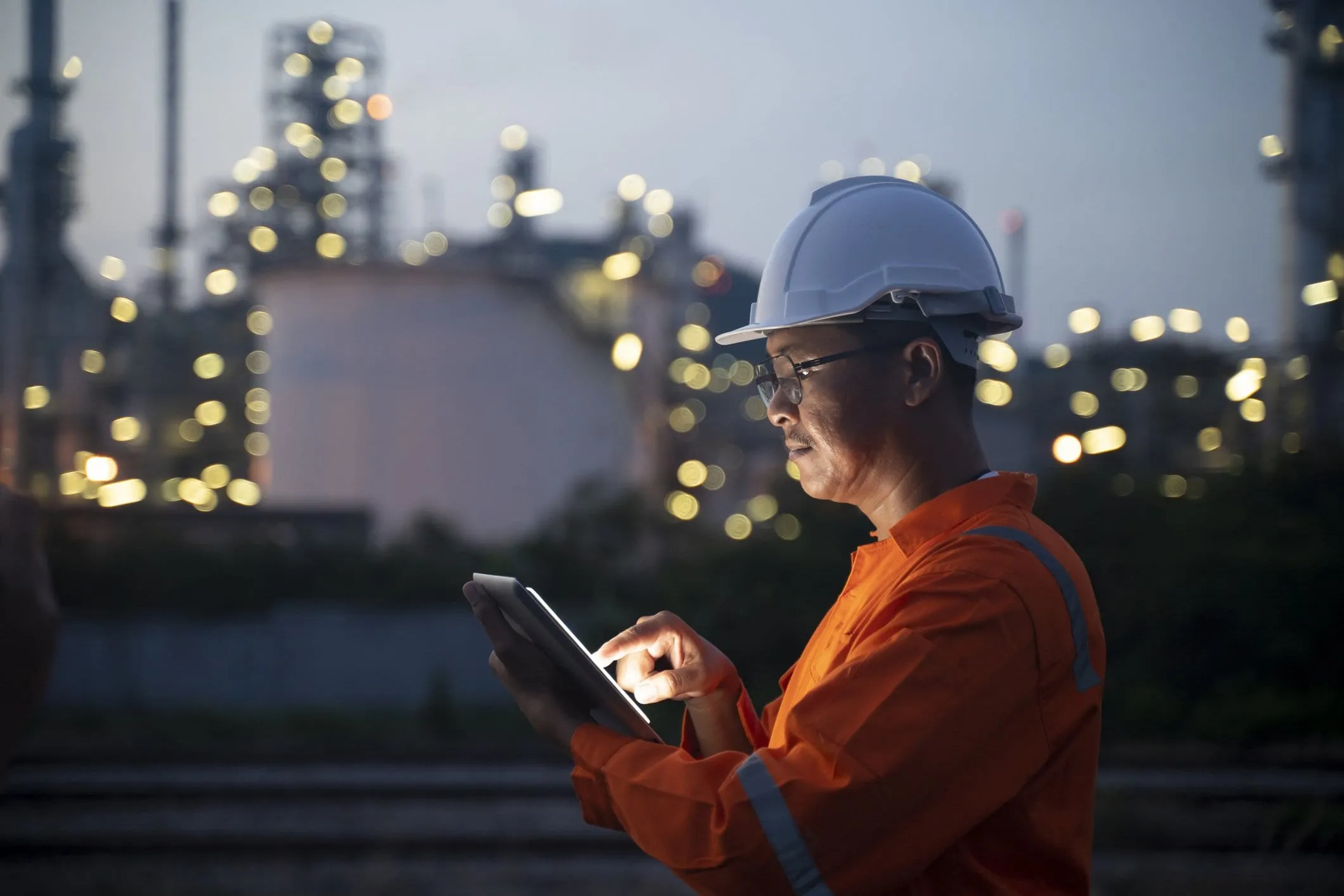 Ein Servicetechniker mit Helm bedient in der Abenddämmerung ein Tablet vor einer Industrieanlage
